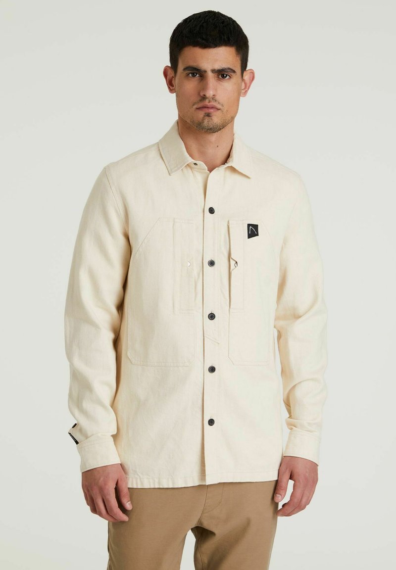 Camicia button-up di colore beige chiaro con maniche lunghe, due tasche frontali, bottoni neri e un piccolo logo nero sulla tasca del petto.