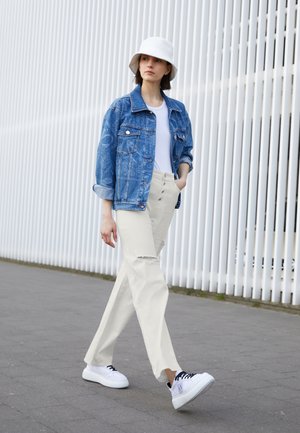 Modelo a usar uma jaqueta de denim azul, t-shirt branca, calças bege claro com um rasgado e tênis brancos com cadarços pretos; em pé num ambiente urbano.