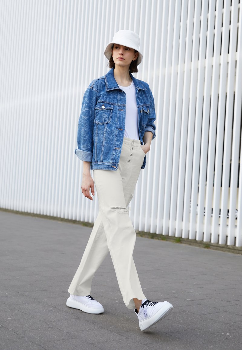 Modelo a usar uma jaqueta de denim azul, t-shirt branca, calças bege claro com um rasgado e tênis brancos com cadarços pretos; em pé num ambiente urbano.