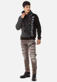 Grauer und schwarzer Kapuzenpullover mit grafischem Design und Logo, kombiniert mit verwaschenen hellgrauen Jeans mit Rissakzenten. Schwarze Schnürstiefel.