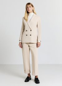 Blazer beige à double boutonnage avec des boutons foncés, associé à un pantalon beige à revers. Porté sur un col roulé blanc, complété par des mocassins noirs.