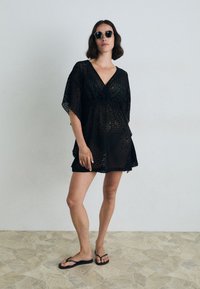 Copertura in pizzo nero con scollatura a V, maniche corte e vita cintrata. Indossata sopra ciabatte nere con un design a motivi.