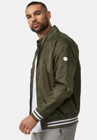 Giacca bomber verde oliva con una texture liscia e lucida; presenta polsini a coste a righe bianche e un logo circolare sul braccio sinistro. Chiusura con zip anteriore.
