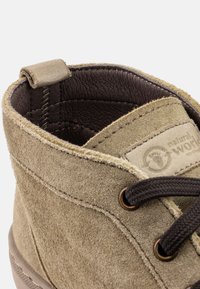 Beige suede bokacsizma barna bőr béléssel. Barna fűzőkkel, saroknál egy hurokkal és logós folttal rendelkezik a nyelvén. Sima textúra.