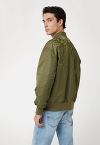 Jonge man met donker haar, die een olijfgroene bomberjack met geborduurde bloemdetails en lichtblauwe jeans draagt, terwijl hij over zijn schouder kijkt.