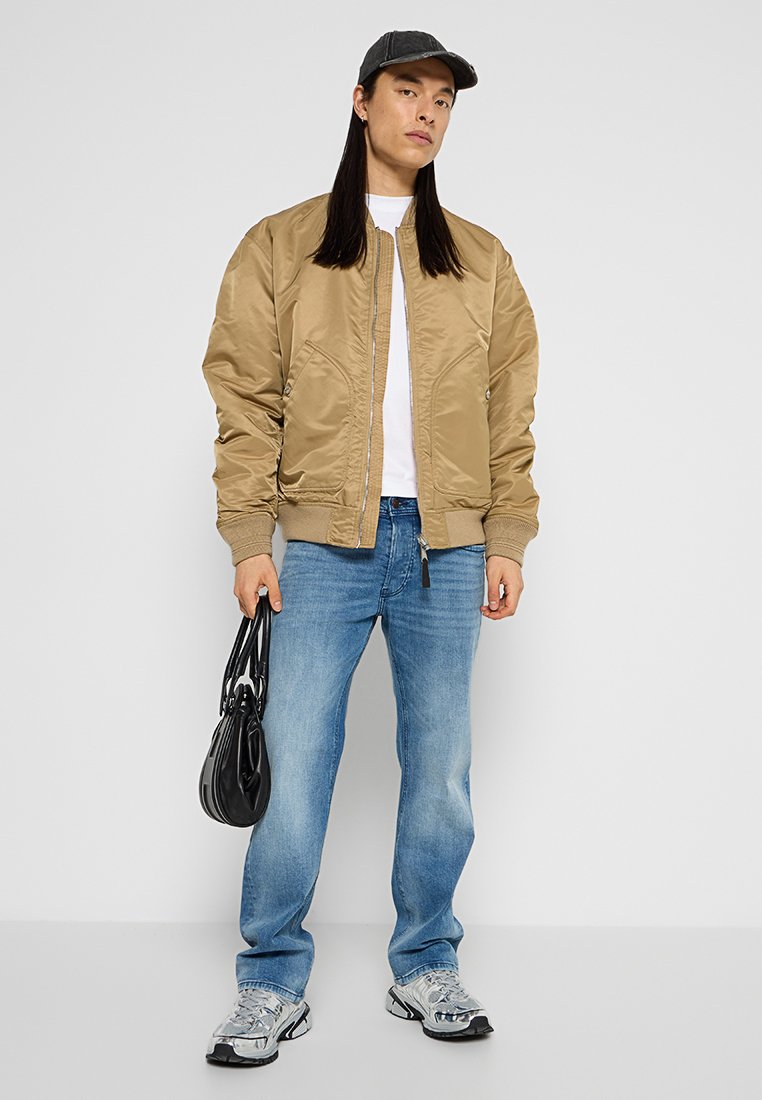 Person mit langen dunklen Haaren, die eine beigefarbene Bomberjacke, ein weißes Shirt, blaue Jeans, graue Sneaker und eine schwarze Kappe trägt, während sie eine schwarze Handtasche hält.