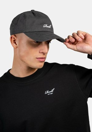 Black cotton cap with a curved visor, featuring white embroidered logo on the front. Model wears a black sweatshirt with a similar logo.