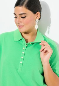 Zielona koszulka polo z kołnierzykiem i trzema guzikami, wykonana z teksturowanej tkaniny. Towarzyszą jej duże, dekoracyjne kolczyki.