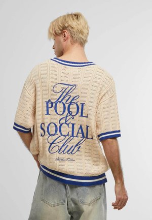 Kootud beež sviiter siniste sooniku aktsentidega, millel on avatud kudumismuster ja suur sinine graafiline tekst "The POOL & SOCIAL Club."