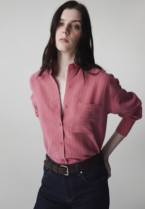Młoda kobieta o długich ciemnych włosach, ubrana w różową koszulę zapiętą na guziki, włożoną w ciemne dżinsy z brązowym paskiem, stoi z jedną ręką za plecami.