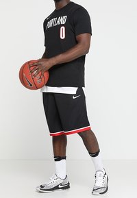 Svart baskettröja med "PORTLAND" och nummer "0" i vitt. Svarta meshshorts med röd trim. Nike-logotyp på tröjan och skorna. Basketboll i handen.