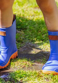 Blaue Gummistiefel mit orangen Sohlen, die über eine strukturierte Oberfläche und eine braune Lasche an der Seite verfügen und in feuchtem, grasigem Gelände getragen werden.
