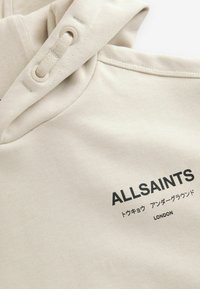 Bež sweatshirt s silediga tekstuuriga, millel on nööriga kapuuts ja musta värvi trükitud tekst "ALLSAINTS", koos lisatekstiga jaapanikeeles.