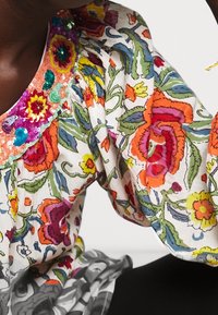 Barevná květinová blůza s bílým pozadím, živé vzory v červené, oranžové a zelené barvě, s výstřihem ozdobeným flitry.