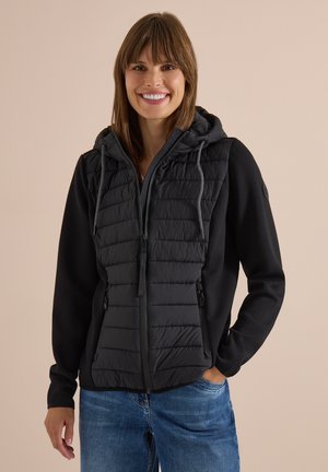 Kvinde smilende iført en sort quiltet hættejakke med lynlås og blå jeans, stående mod en ensfarvet beige baggrund.