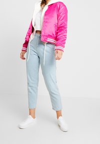 Bright pink satin bomber jacket with white accents, featuring ribbed cuffs and hem, paired with light blue denim jeans and white sneakers.