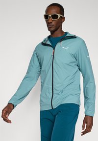 Ελαφρύ, τιρκουάζ μπουφάν εξωτερικού χώρου με κουκούλα, πλήρες φερμουάρ και μαύρες λεπτομέρειες. Διαθέτει πορτοκαλί τράβηγμα και μια διακριτική λογότυπο στο στήθος.