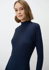 Donker marineblauwe ribgebreide jurk met een hoge kraag, voorzien van een chevronpatroon en lange mouwen, die een lichaamsomsluitende silhouet toont.