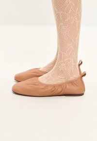 Bailarinas de cuero marrón claro con punta redondeada, diseño fruncido y pequeño detalle de lazo en la parte trasera, combinadas con medias de red color nude.