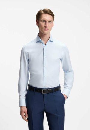 Mann trägt hellblaues Hemd, das in dunkelblaue Hose mit schwarzem Gürtel gesteckt ist, steht mit einer Hand in der Tasche vor weißem Hintergrund.