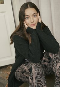 O tânără cu păr lung și castaniu, așezată pe podea, purtând un pulover negru și pantaloni cu model, își odihnește fața în palme.