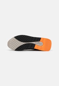 Schwarze Sneaker-Sohle mit einer strukturierten, rutschfesten Oberfläche. Mit grauen und orangefarbenen Akzenten. Beinhaltet ein auffälliges Logo und ein gemustertes Detail.
