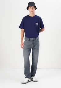 Marineblauwe katoenen t-shirt met witte logo, gecombineerd met lichtblauwe denim jeans. Het model draagt een zwarte bucket hat en witte sneakers met zwarte strepen.