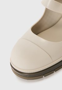 Beige kummist Mary Jane kingad ümmarguse varbaga, laia rihmaga, tekstuuriga sisetalla ja kontrastselt tumeda tallaga, mille serv on mustriline.