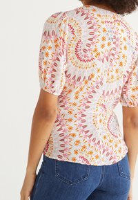 Blusa bianca con motivo floreale multicolore, maniche a sbuffo e vita aderente, abbinata a jeans blu scuro.