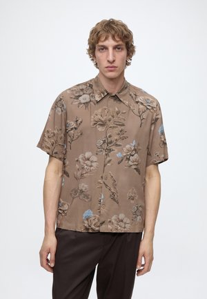 Jeune homme portant une chemise marron à manches courtes avec des motifs floraux beige et bleu clair et un pantalon foncé, debout devant un fond blanc uni.
