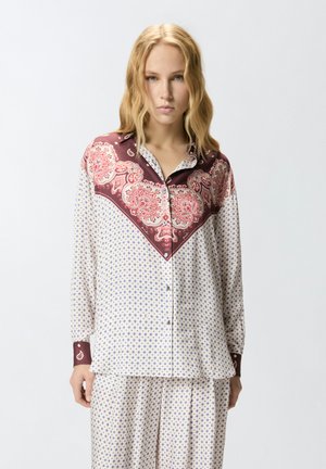 Mujer rubia vistiendo un conjunto suelto de camisa blanca y pantalones con pequeños patrones azules y un diseño de bandana de estilo cachemira en color granate encima.