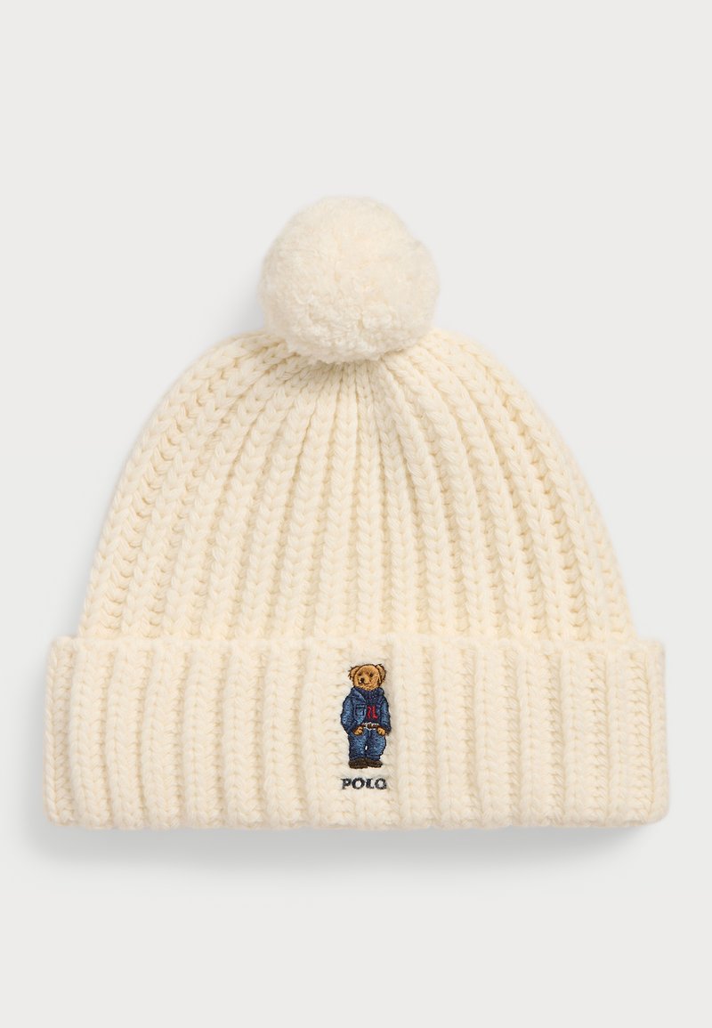 Gorro de malha creme com um padrão texturizado, featuring um pompom no topo e um urso bordado a usar um fato azul com o texto "Polo".
