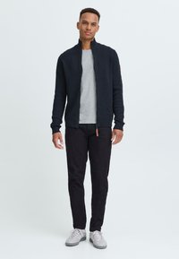 Cárdigan azul marino con cremallera y puños acanalados, llevado sobre una camiseta gris clara. Combinado con pantalones negros y zapatillas grises. Diseño simple y ajustado.