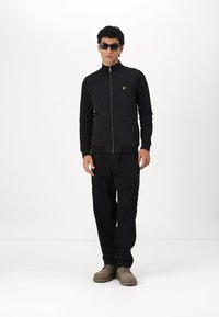 Giacca nera con zip e colletto alto, pannello frontale testurizzato e logo giallo. Abbinata a pantaloni cargo neri e stivali beige. Occhiali da sole indossati.