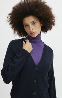 Cardigan bleu marine en tissu doux et texturé avec un décolleté en V, comportant quatre boutons assortis. Porté par-dessus un col roulé violet.