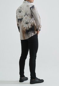 Chemise à manches longues présentant un motif floral dans des tons beiges et noirs, fabriquée en tissu léger avec un col à boutons et des poignets.