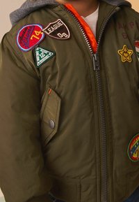 Blouson aviateur vert olive avec une capuche grise, agrémenté de plusieurs patchs colorés et une doublure orange ; fermeture éclair à l'avant et poche latérale zippée.