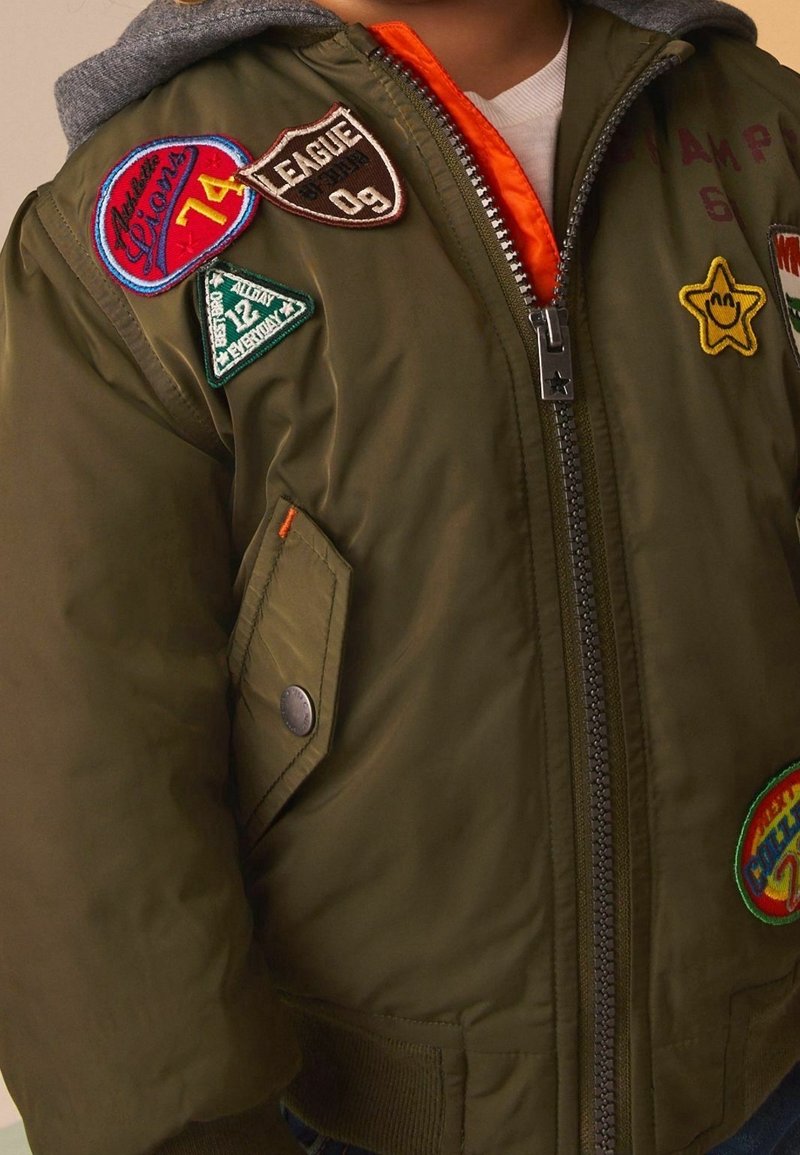 Blouson aviateur vert olive avec une capuche grise, agrémenté de plusieurs patchs colorés et une doublure orange ; fermeture éclair à l'avant et poche latérale zippée.