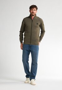 Olivgrüner Reißverschluss-Pullover mit gerippten Akzenten, kombiniert mit hellblauen Jeans und weißen Sneakers. Lässige Passform, strukturierter Stoff und dezente Logodetails.