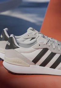 Baskets blanches Adidas avec des bandes noires et un détail au talon, placées sur une surface orange avec un fond bleu sous une lumière douce.