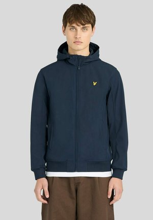 Jonge man die een marineblauwe zip-up hoodie met capuchon en klein geel logo op de linkerborst draagt, bruine broek, staand tegen een effen achtergrond.