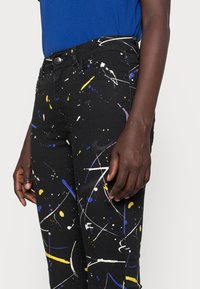 Jeans negros con salpicaduras de pintura multicolor en amarillo, azul y blanco, con un corte clásico de denim y un cierre de botón en la cintura.