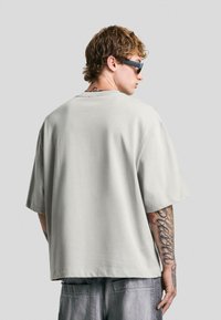 Mladý muž s kudrnatými vlasy a tetováním nosí oversized světle šedé tričko, tmavé sluneční brýle a volné džíny, stojí bokem proti jednobarevnému pozadí.