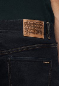 Σκούρο τζιν με πορτοκαλί ραφές, δερμάτινο ταμπελάκι με στυλιζαρισμένη φιγούρα και αστέρια, και μικρό λογότυπο «VOLCOM» στην πίσω τσέπη.