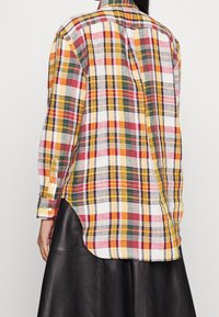 Colorful plaid long-sleeve shirt with red, yellow, green, and white patterns worn over a black leather skirt, shown from the back.