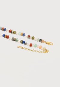 Collier de perles en pierre multicolores et perles avec rallonge de chaîne en or et fermoir mousqueton sur fond blanc.