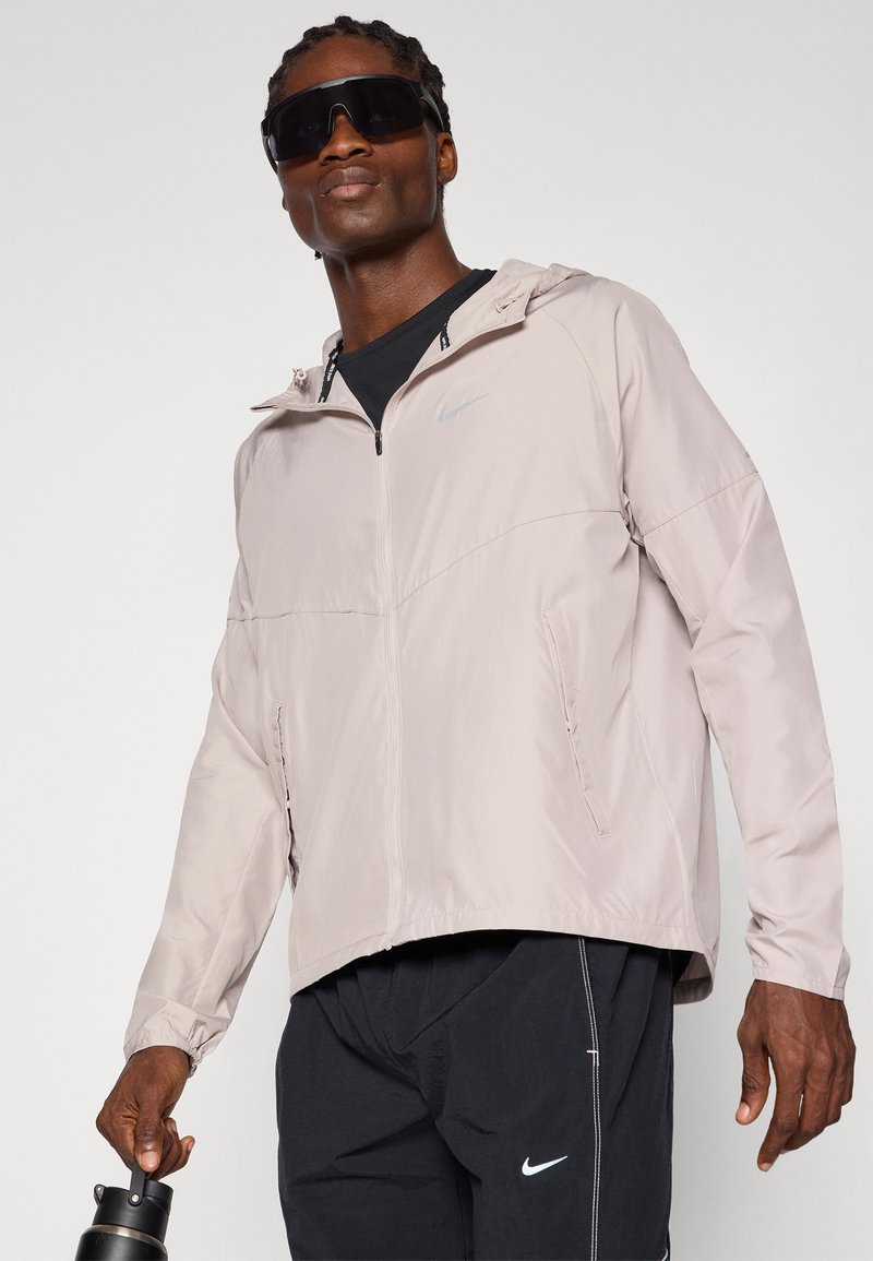 Helle beige Windbreaker-Jacke mit Reißverschluss vorne, Kapuze und elastischen Bündchen. Ausgestattet mit dezentem Nike-Logo und Seitentaschen. Getragen über einem schwarzen Hemd.