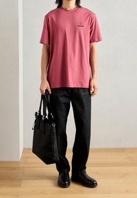Camiseta de algodón rosa con un logotipo gráfico negro, combinada con pantalones de mezclilla oscuros y una bolsa tote negra que presenta múltiples bolsillos y correas.