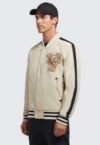 Beige bomberjakke med en sort og beige stripet ribkant. Har et broderet pantherlogo og sidelommer. Suppleret med en sort kasket.