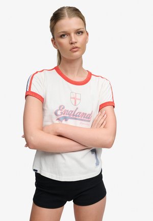 Jeune femme aux cheveux blonds, bras croisés, portant un maillot blanc de l’Angleterre avec bordure rouge et un short noir, debout devant un fond uni.